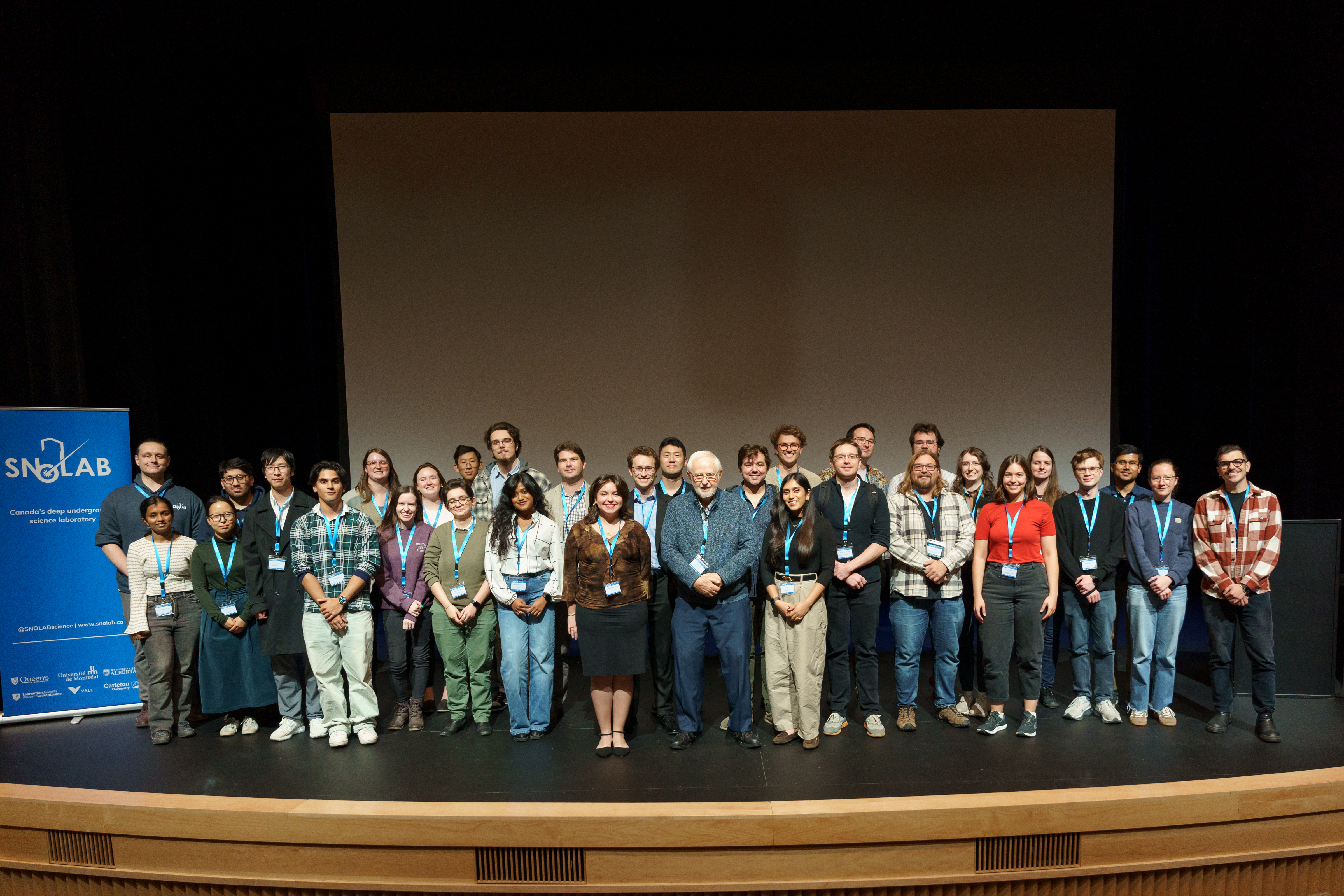 Highly qualified personnel (student, post-doctoral researcherc, and other early career folks) stand on stage with 2015 Nobel Laureate Dr. Art McDonald